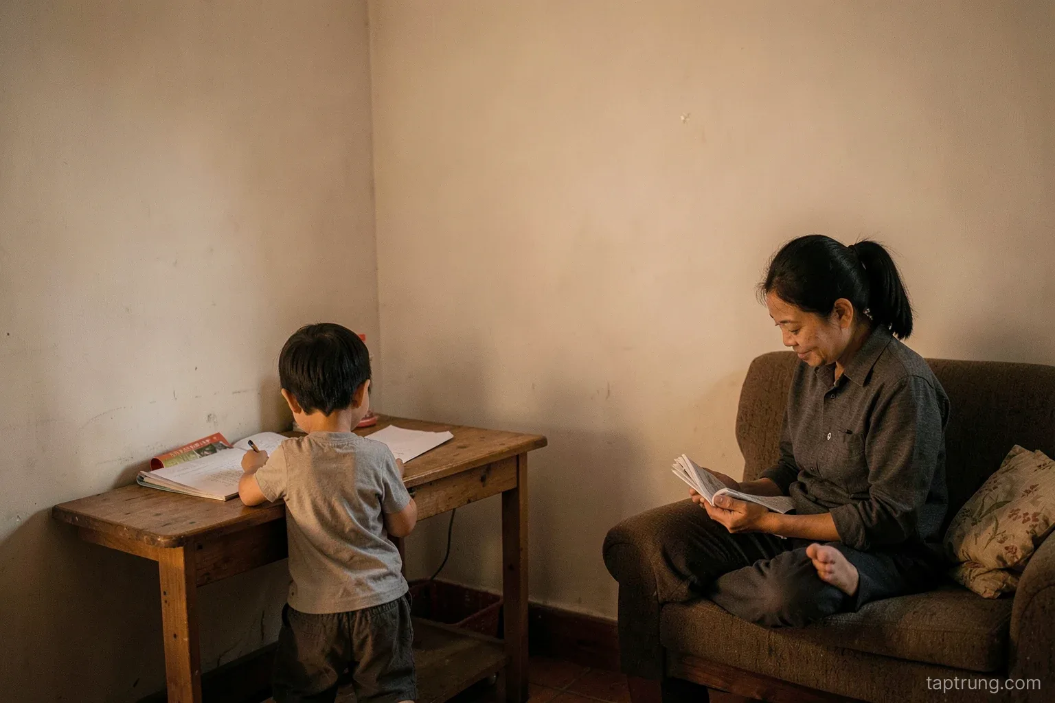 Wide shot of a quiet corner in a small Vietnamese apartment, simple desk facing a plain pale wall, child seen from behind writing, parent reading on a sofa nearby, warm evening light